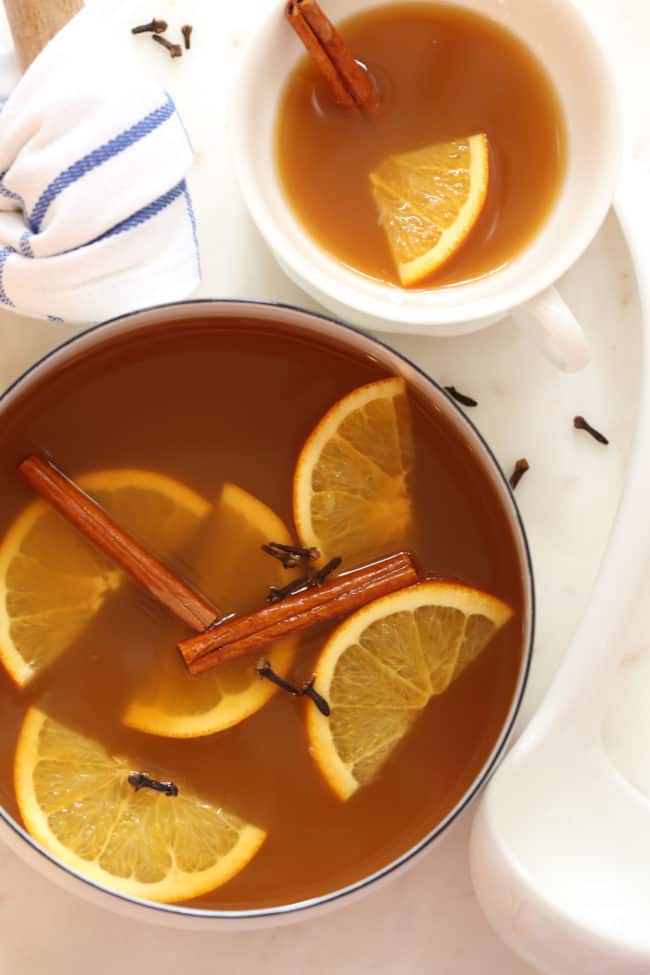A white pot filled with a hot drink with orange slices and cinnamon sticks.