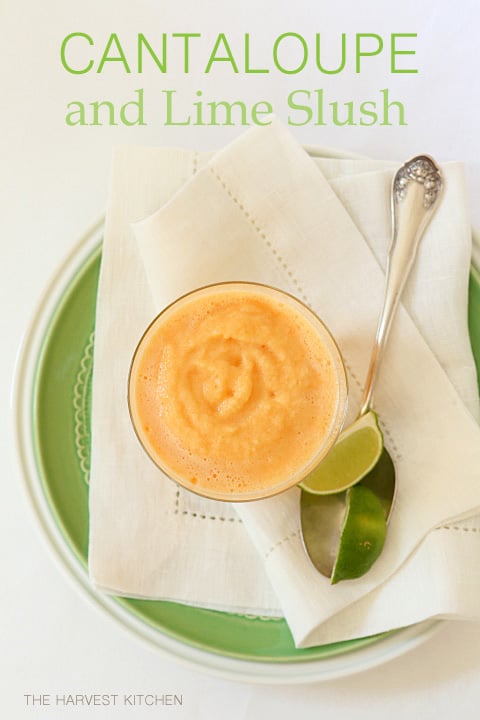 A clear drinking glass filled with cantaloupe slush. The glass sits on a green plate next to a spoon.