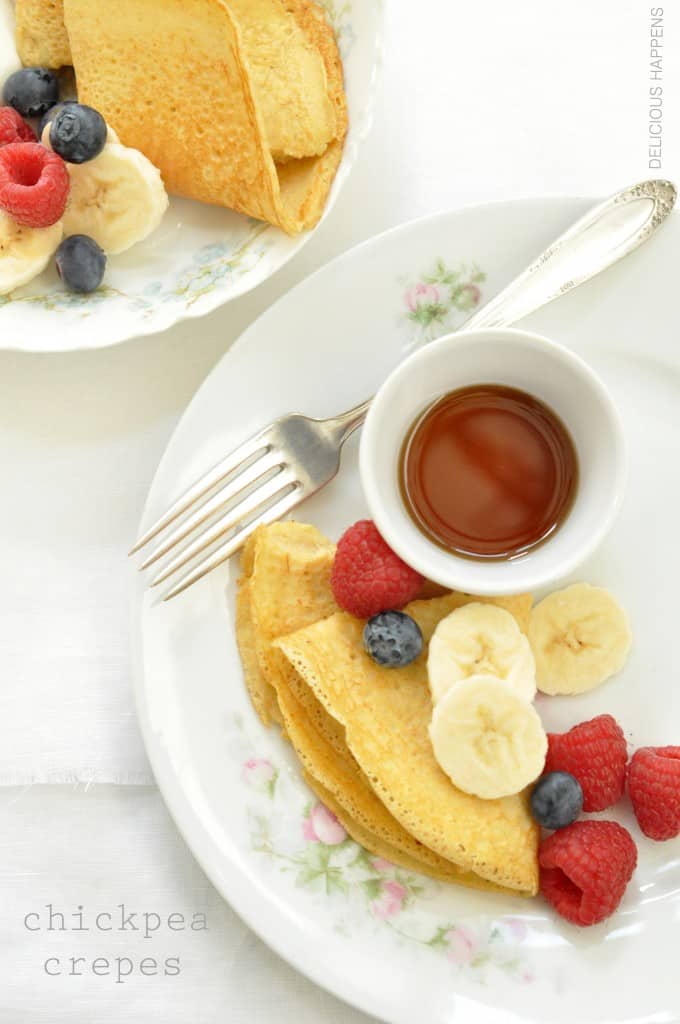 A white plate with chickpea crepes and fresh fruit and a small white bowl filled with pure maple syrup.