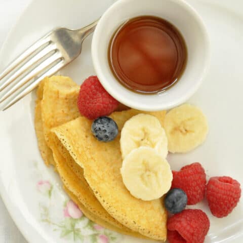 A white plate with chickpea crepes, fresh fruit and a small bowl filled with pure maple syrup.