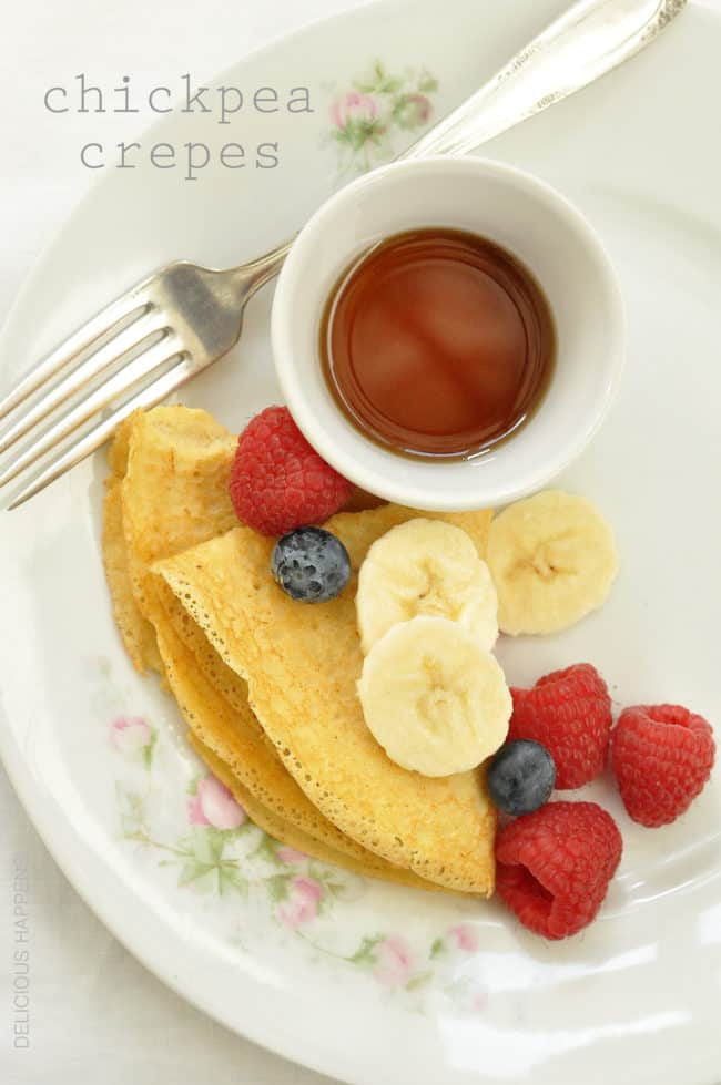 A white plate with chickpea crepes, fresh fruit and a small white bowl of pure maple syrup.