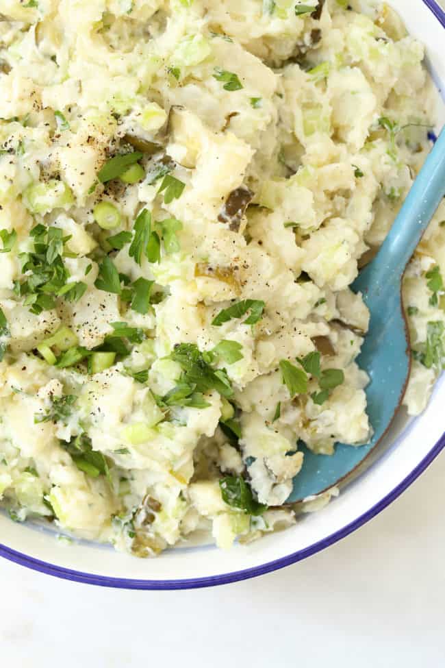 A white bowl filled with Dill Pickle Potato Salad. A blue serving spoon rests in the bowl.