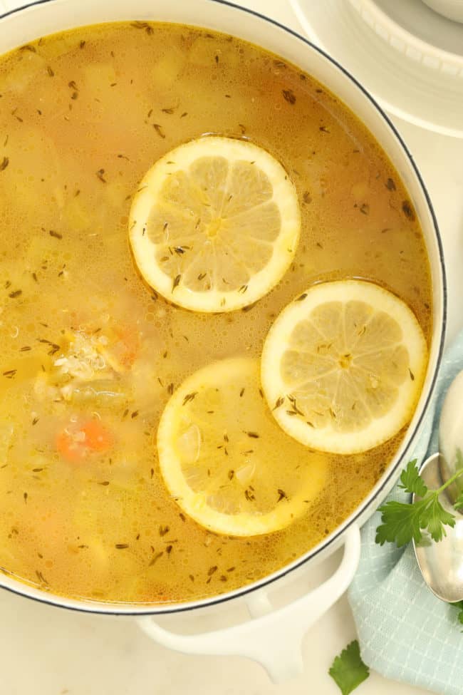 A white pot filled with lemon chicken soup. Three slices of lemon float on top of the soup.