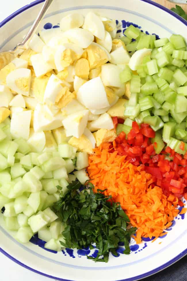 A white bowl filled with chopped hardboiled egg, celery, cucumber, parsley and grated carrot.