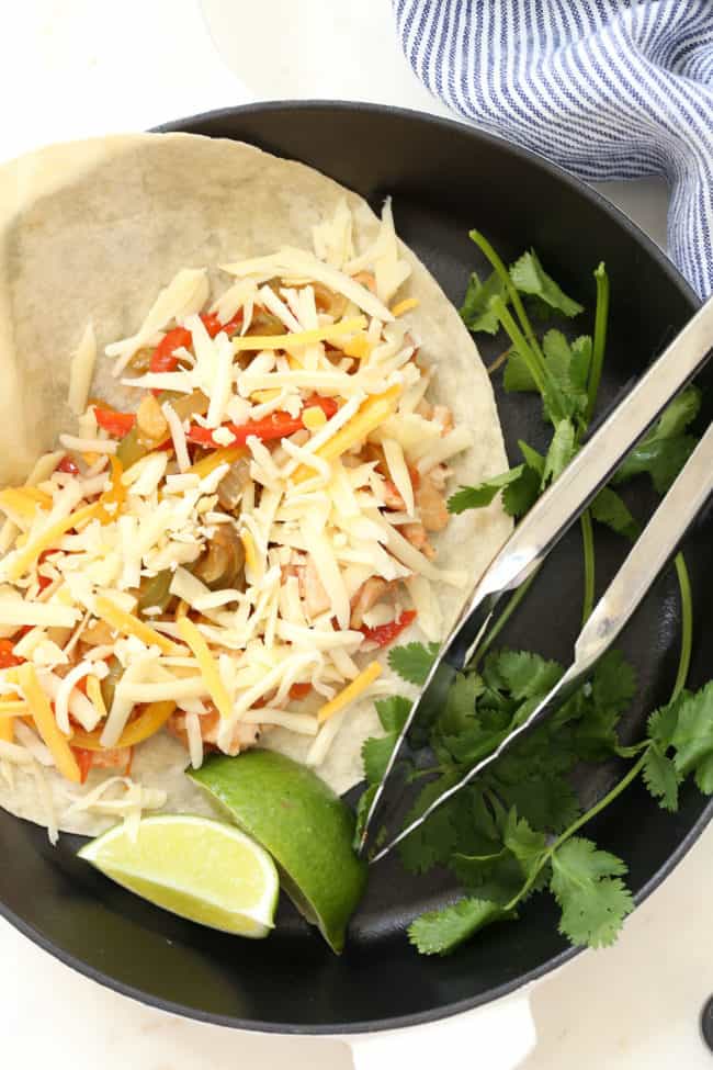 A black cast iron skillet with cooked poultry strips and vegetables on a flour tortilla with grated cheese. 