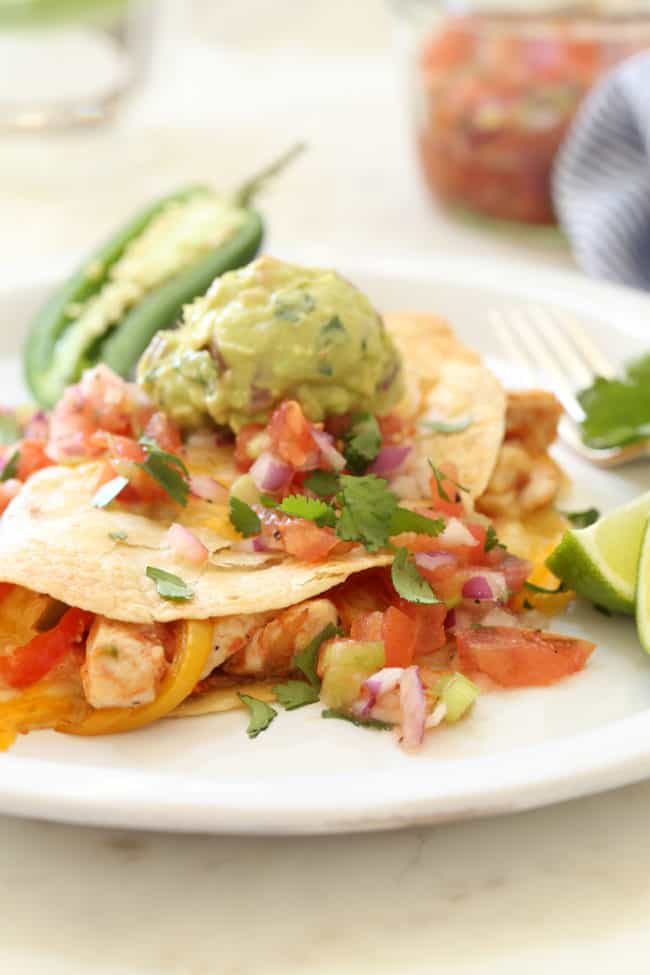 A white plate with a baked chicken fajita quesadilla on it. The quesadilla has a scoop of guacamole and pico de gallo on top of it. Recipe for baked quesadillas.