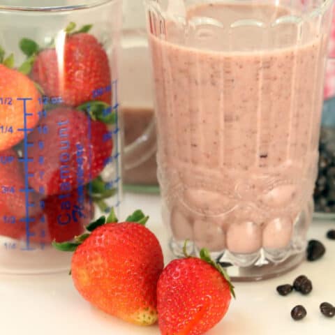 A clear glass filled with strawberry cacao nibs smoothie. Strawberries and cacao nibs are scattered next to the glass.