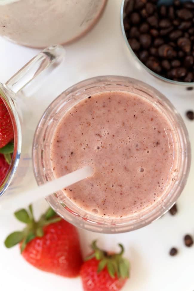 a clear glass filled with strawberry cacao nibs smoothie. Strawberries and cacao nibs are scattered next to the glass.