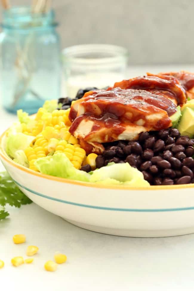A bowl filled with lettuce, black beans, corn, avocado, grated cheddar cheese and BBQ chicken