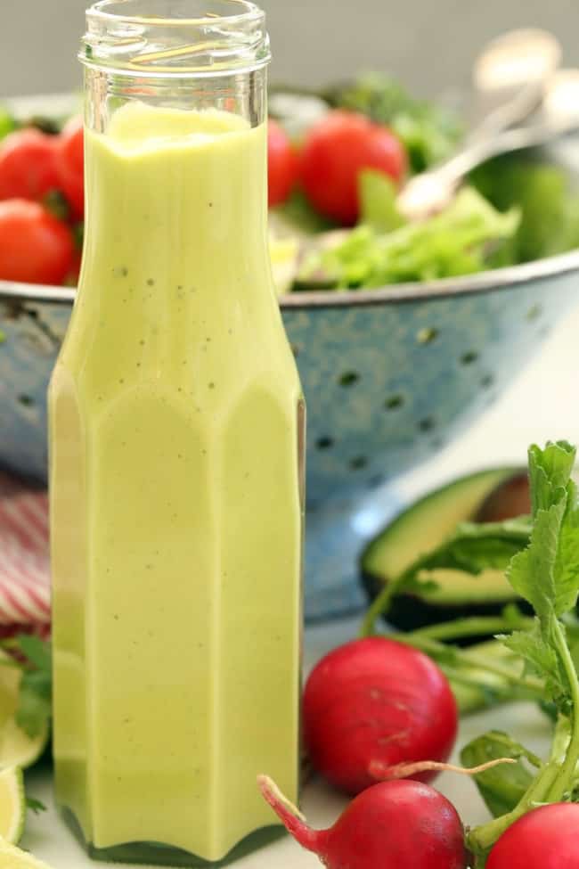 A clear bottle filled with avocado salad dressing, with a blue colander behind bottle filled with salad.