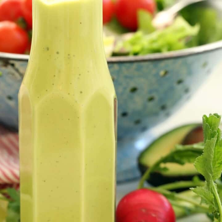 A clear bottle filled with avocado salad dressing. Blue colander behind bottle filled with salad.