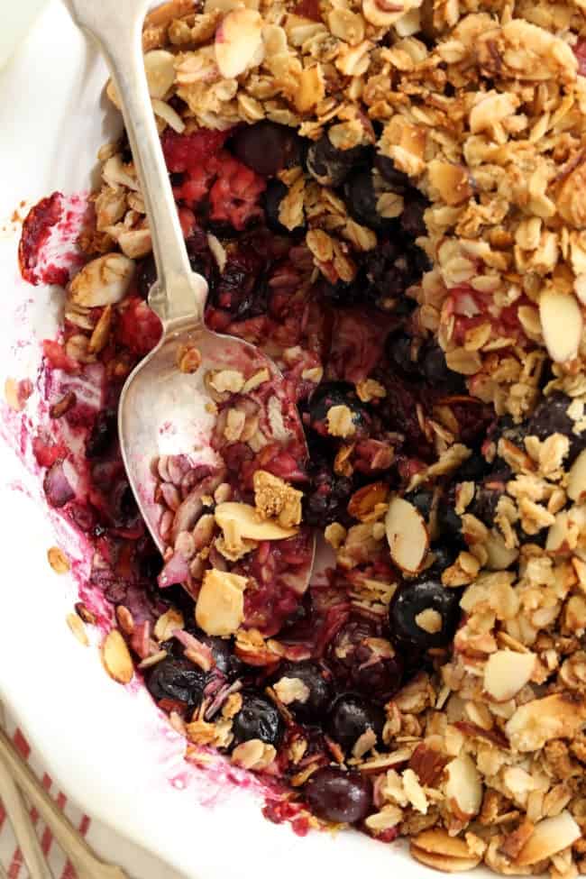 A white pie dish filled with triple berry crisp. A silver spoon rests in the dish.