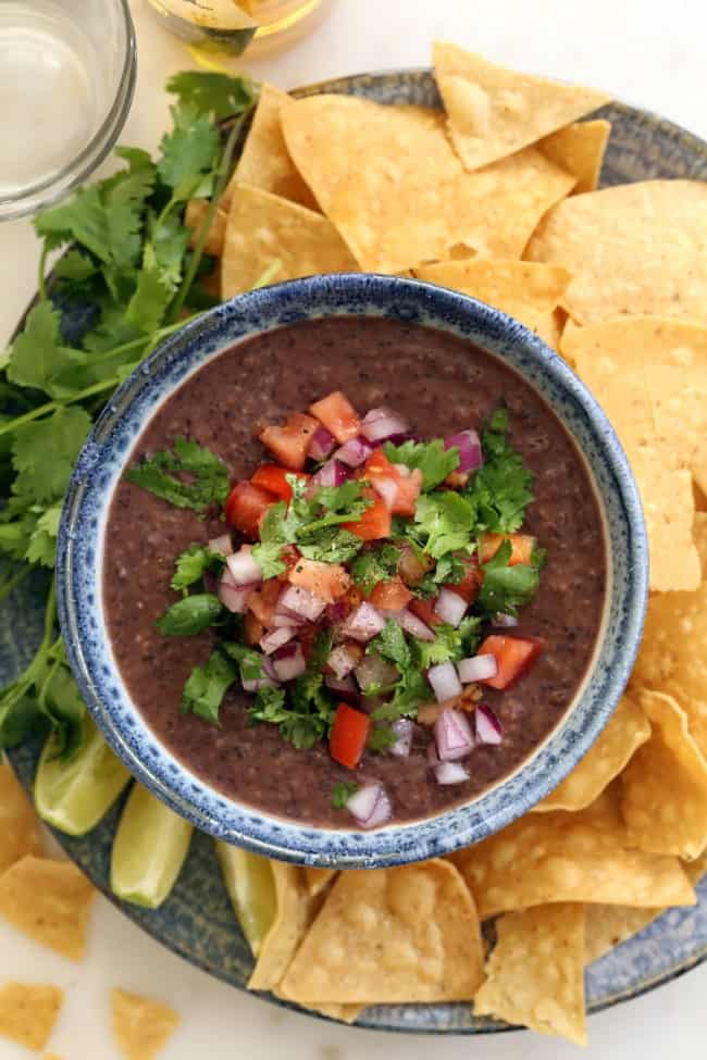 A blue-rimmed bowl filled with mashed legumes and salsa on a plate filled with tortilla chips.