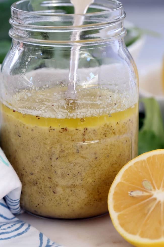 A glass mason jar half filled with lemon mustard vinaigrette. A blue and white napkin and half a lemon sits next to the jar.