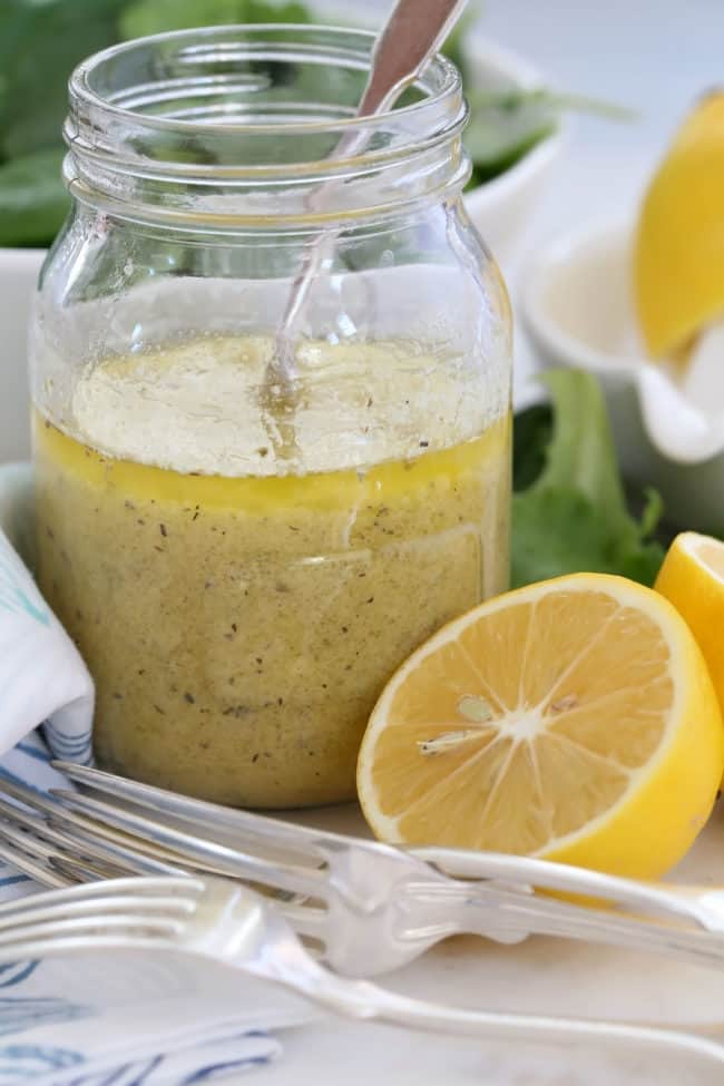 A glass mason jar half filled with homemade salad dressing. A blue and white napkin, forks and half a lemon sits next to the jar.