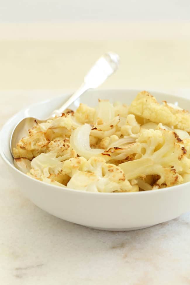 A white bowl filled with roasted cauliflower. A serving spoon rests in the bowl.