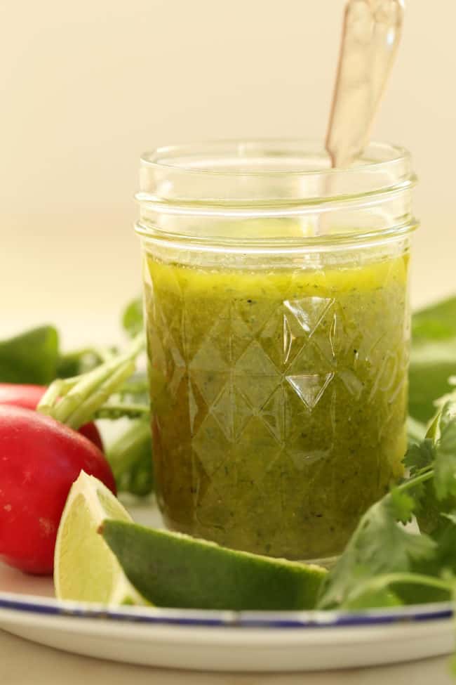 clear glass mason jar filled with cilantro lime vinaigrette. The jar sits on a white plate with radishes, limes and cilantro.