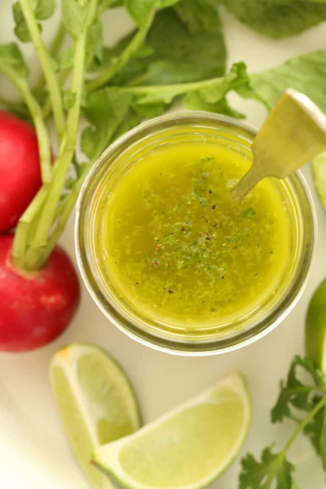 clear glass mason jar filled with a healthy salad dressing with cilantro. The jar sits on a white plate with radishes, limes and cilantro.