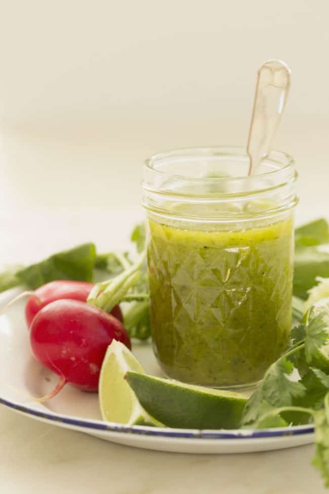 clear glass mason jar filled with cilantro lime vinaigrette. The jar sits on a white plate with radishes, limes and cilantro.