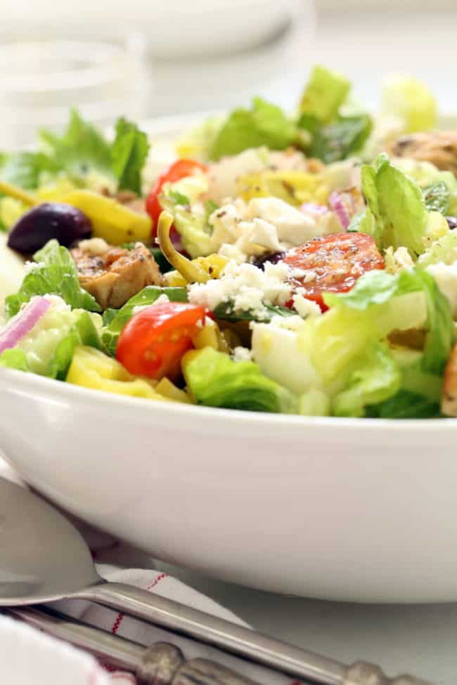 White bowl filled with salad and grilled chicken. Silver serving spoons sit next to the bowl.