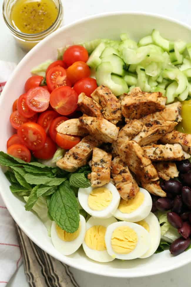White bowl filled with grilled chicken, tomatoes, cucumbers, kalamata olives and Romaine lettuce.