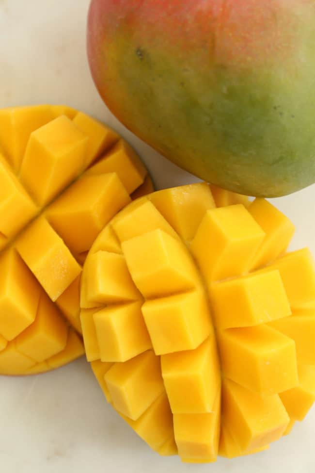 Two mangoes on a white background. One mango is cut into cubes.