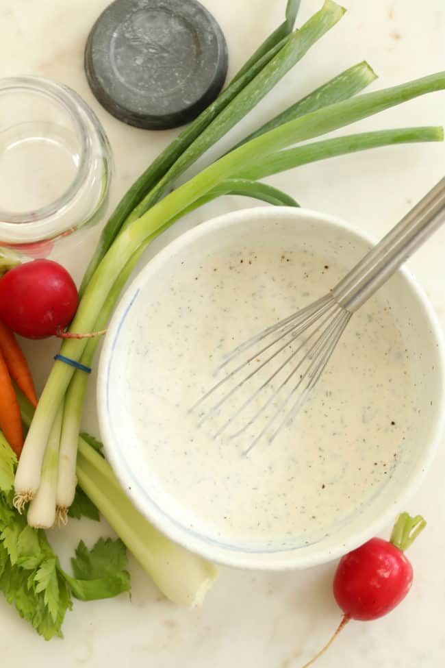 white bowl filled with ranch dressing. A silver whisk is in the bowl and carrots, celery, green onion and radishes lay next to the bowl