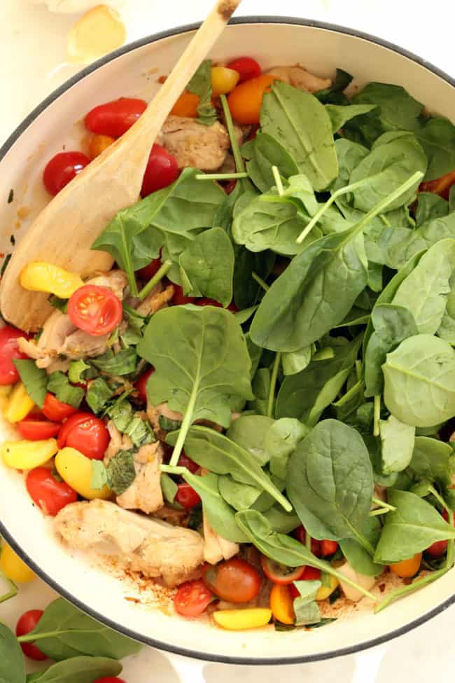 A white pan filled with cooked poultry, cherry tomatoes and raw spinach. A wooden spoon rests in the pan.