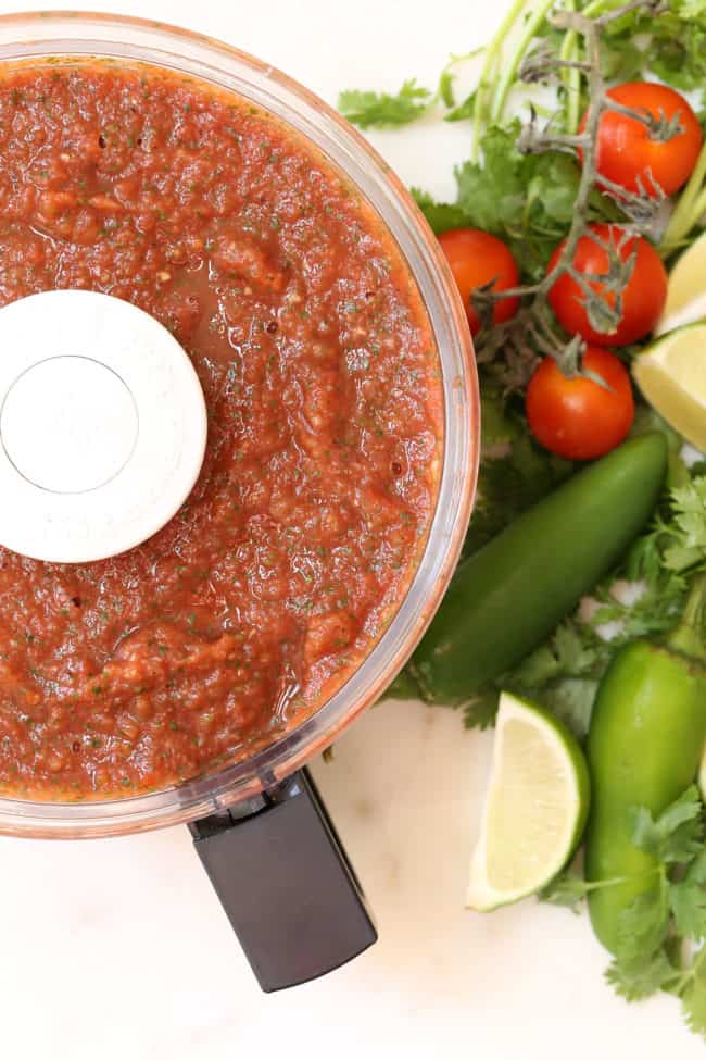 A food processor filled with restaurant style salsa. Cherry tomatoes and jalapeno pepper sit next to the processor.