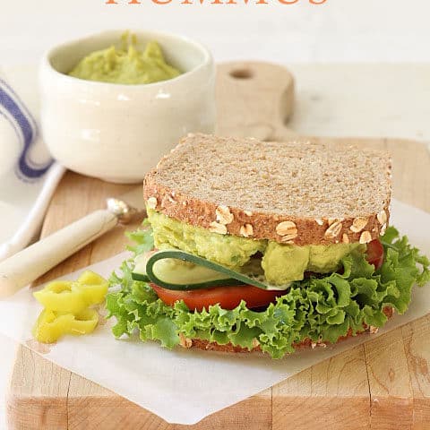A sandwich made with Avocado Hummus, lettuce, cucumber and tomato sits on a wooden cutting board.