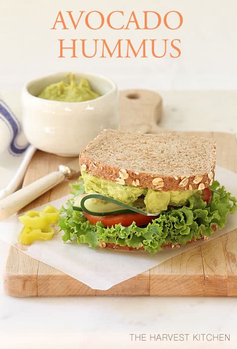 A sandwich made with Avocado Hummus, lettuce, cucumber and tomato sits on a wooden cutting board.