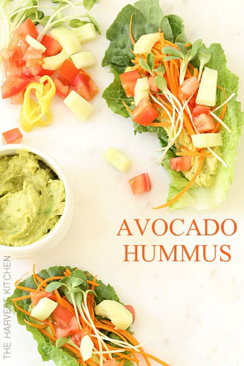 A small white bowl filled with Avocado hummus. Two lettuce wraps filled with the hummus and vegetables sit next to the bowl.