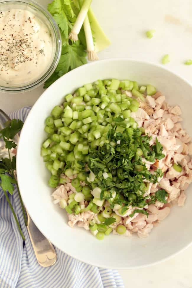 A white bowl of chopped cooked chicken with chopped green onions, celery and parsley.