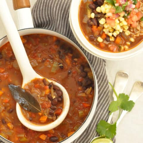 A white pot filled with Black Bean Soup. A ladle rests in the pot and a white bowl filled with the soup sits next to the pot.