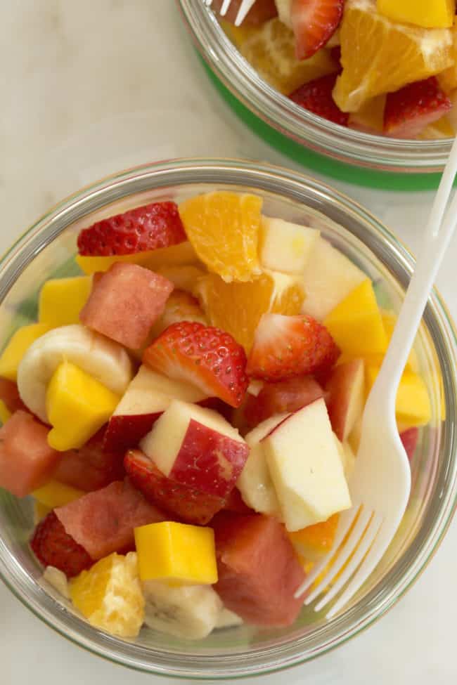 A glass container filled with chopped citrus, bananas and berries. A plastic white fork rests in the container.