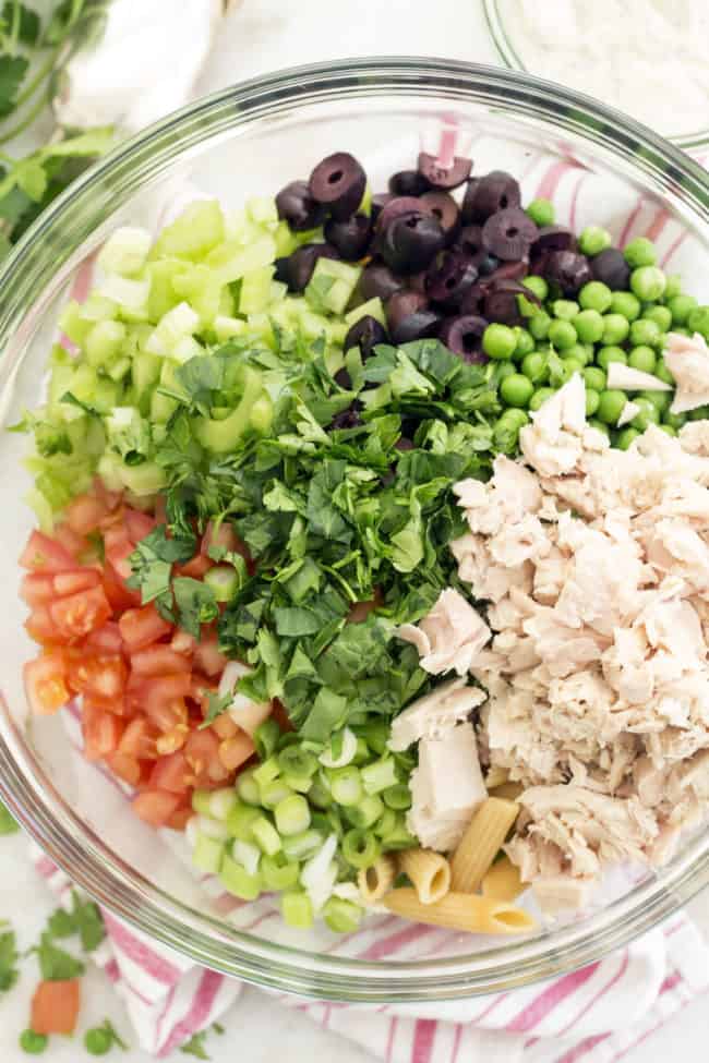 Clear glass bowl filled with canned tuna, celery, tomatoes, green onion, kalamata olives and penne noodles.