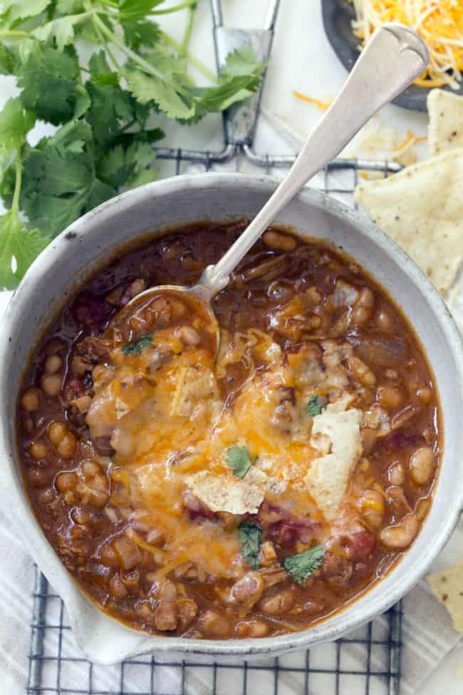 Bowl of white bean turkey chili. A spoon sits in the bowl and sprigs of cilantro, tortilla chips and shredded cheese sit next to the bowl.