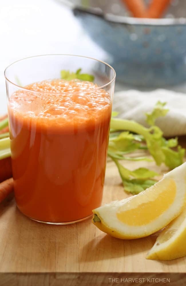 A clear glass filled with carrot juice. Lemon wedges and celery sticks are placed next to the glass. 