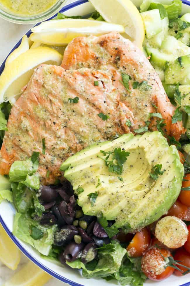 bowl filled with lettuce, grilled salmon, slices of avocado, cherry tomatoes, cucumber and kalamata olives