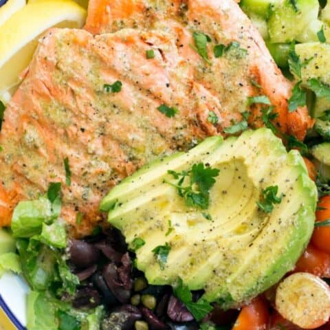 bowl filled with lettuce, grilled salmon, slices of avocado, cherry tomatoes, cucumber and kalamata olives