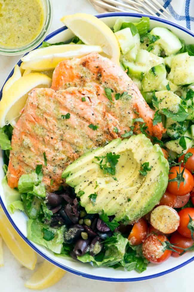 bowl filled with lettuce, grilled salmon, slices of avocado, cherry tomatoes, cucumber and kalamata olives