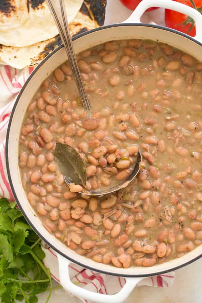 A white pot filled with cooked legumes. A silver ladlel rests in the pot.