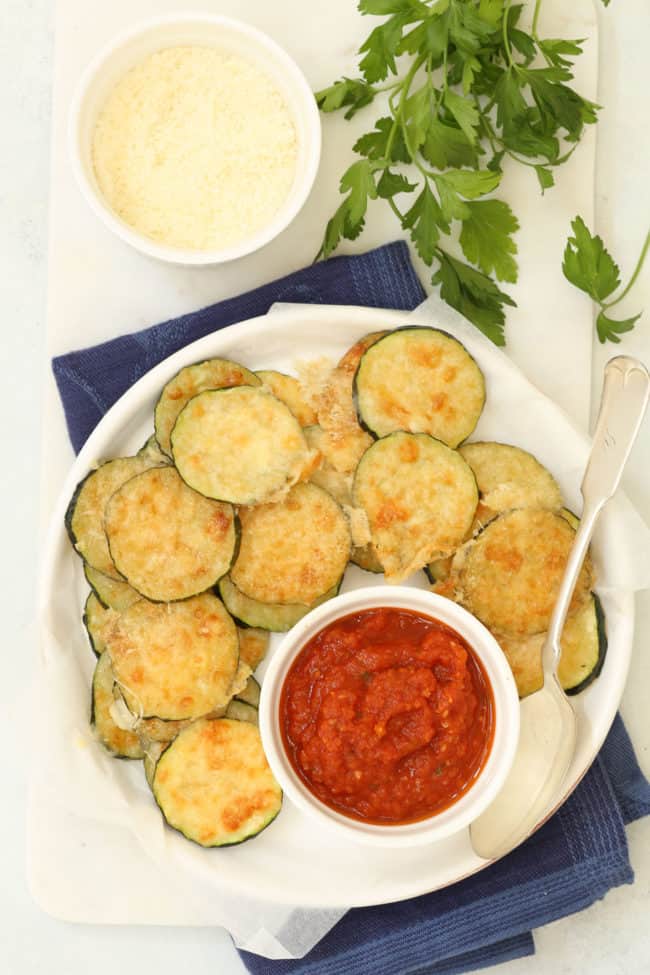 A white serving plate filled with baked zucchini chips. 