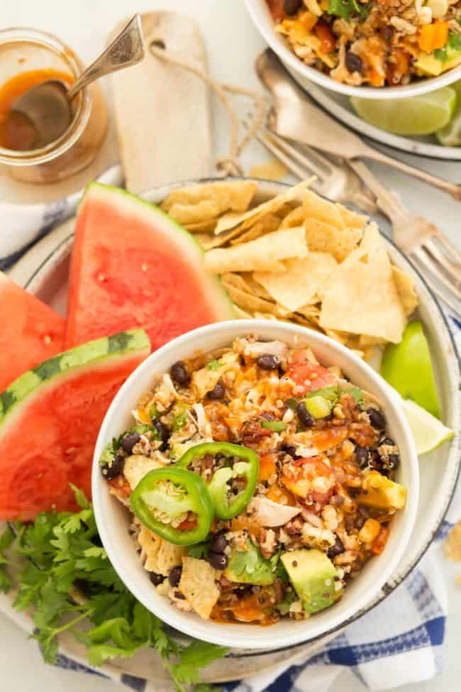 A bowl of chicken, black beans and vegetables on a platter next to tortilla chips and watermelon.