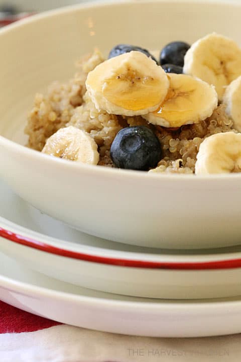 A white bowl of hot oatmeal and quinoa cereal with bananas and blueberries drizzled with honey.