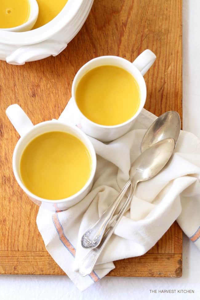 Two white mugs filled with butternut curry soup sits on a wood cutting board. Two soup spoons rest next to the cups.