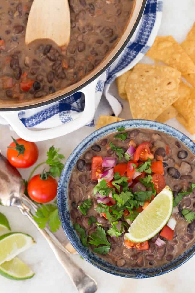 A blue bowl filled with creamy black beans with coconut milk sits next to a large pot of the beans. A wooden spoon rests in the pot.