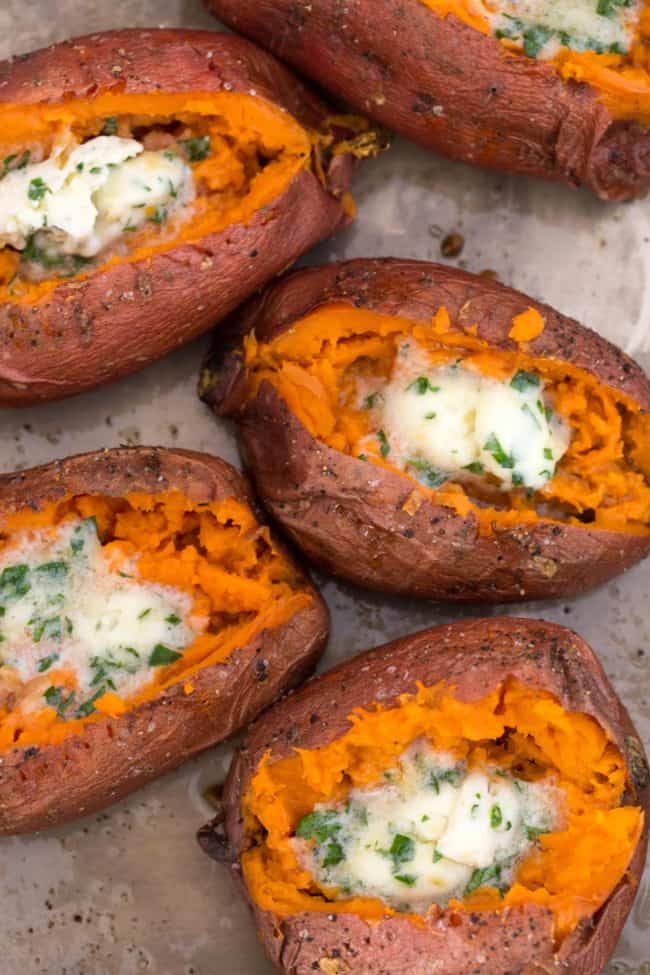 Five baked sweet potatoes stuffed with melting herbed butter on a baking sheet.