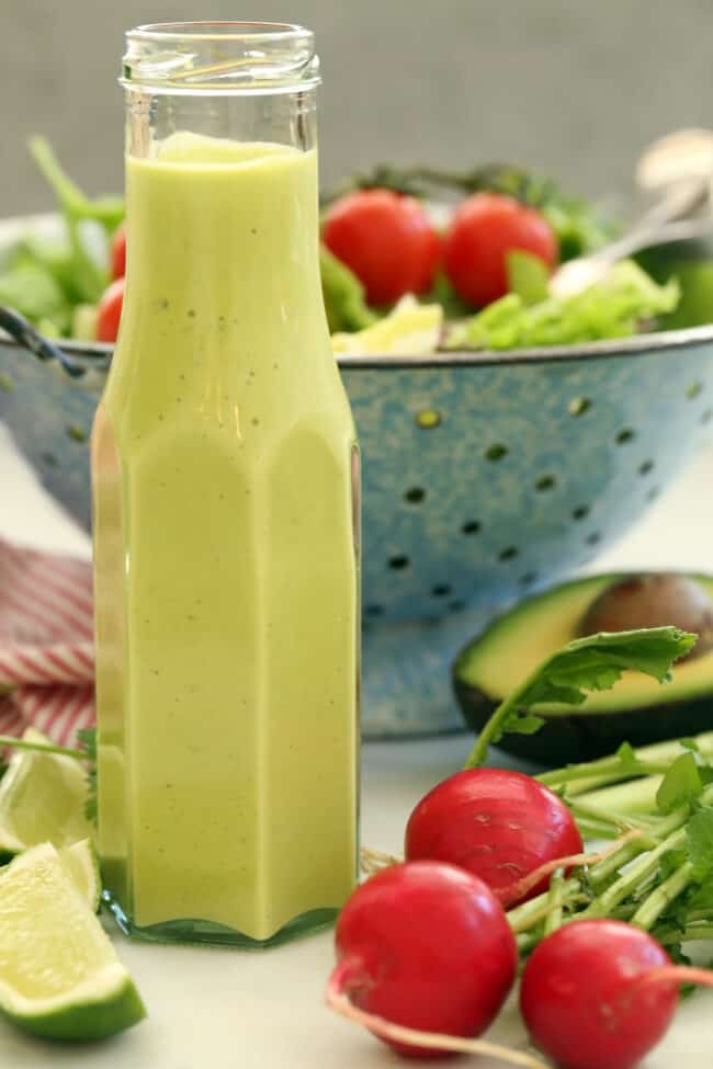 A clear bottle filled with avocado salad dressing. A blue colander behind the bottle filled with salad.