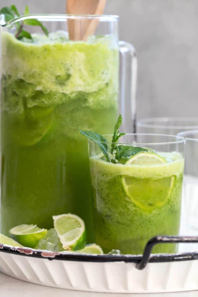 A clear glass next to a lear glass pitcher, both filled with a green drink.